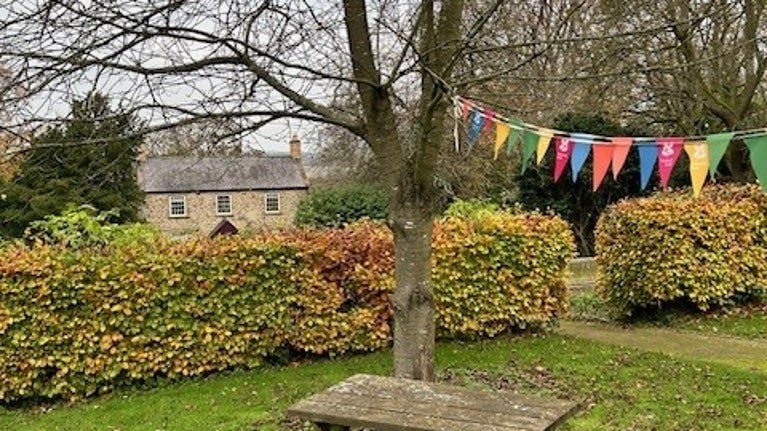A wooden picnic bench on the grass with a tree with colourful bunting behind. The bushes are turning autumn colours and the Cherryburn museum is in the distance
